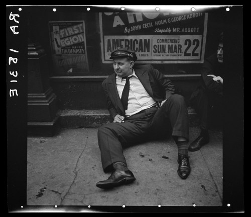 A man in a suit sits on the ground, leaning back against a wall, with vintage posters in the background.