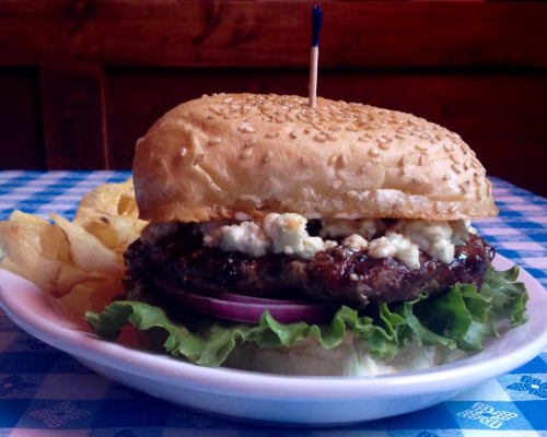 A sesame seed bun burger with lettuce, onion, blue cheese, and a side of potato chips on a blue checkered tablecloth.