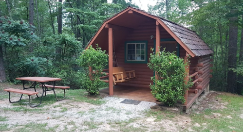 A cozy log cabin surrounded by trees, featuring a porch swing and a picnic table nearby.