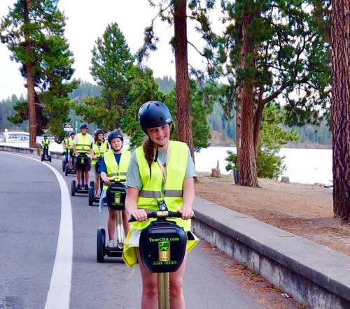 A group of people in bright vests riding segways along a scenic lakeside path surrounded by trees.