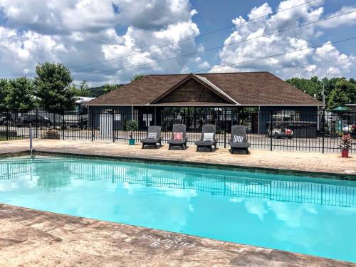 A clear swimming pool with lounge chairs nearby, surrounded by a fence and a building in the background under a cloudy sky.