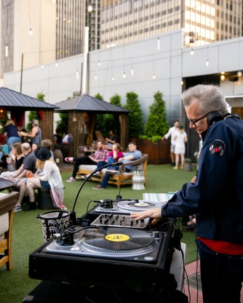 A DJ mixes music on turntables at a rooftop party, with guests socializing in the background.