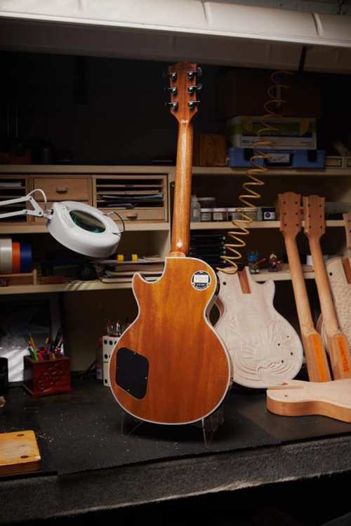 A wooden guitar stands upright on a workbench, surrounded by tools and other guitar parts in a workshop setting.