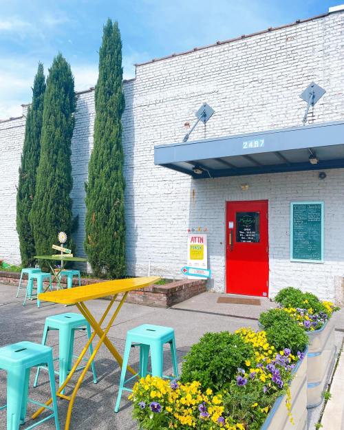 A bright red door on a white brick building, surrounded by green trees and colorful flowers, with outdoor seating.