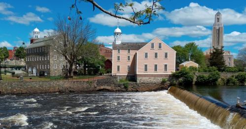 A scenic view of a river with a waterfall, flanked by historic buildings and a blue sky with fluffy clouds.