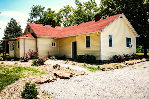 A yellow house with a red roof surrounded by greenery and a gravel pathway.