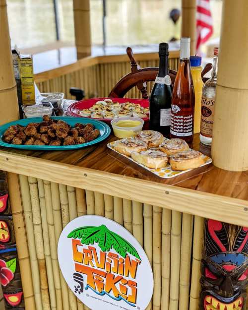 A tiki bar table with snacks, drinks, and a vibrant logo, set against a scenic waterfront backdrop.