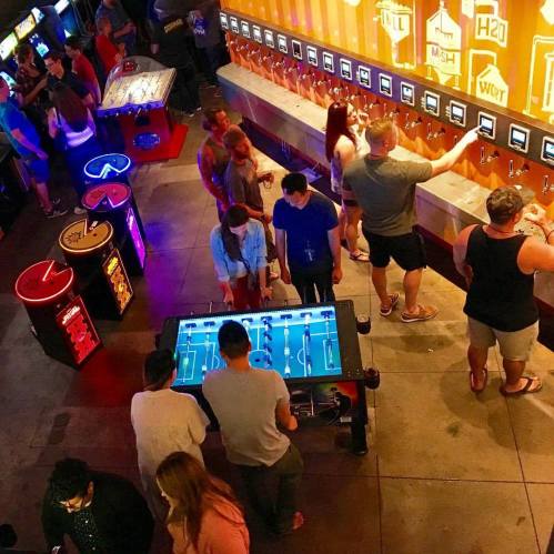 A lively bar scene with people enjoying games and a self-serve drink wall, illuminated by colorful lights.