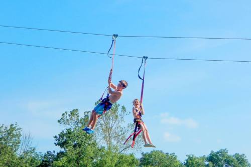Two children ziplining against a clear blue sky, surrounded by trees and enjoying a sunny day outdoors.