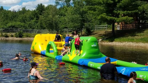Children play on a colorful inflatable water slide in a lake, with adults supervising nearby.