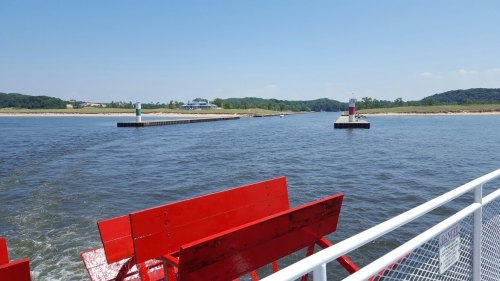 View of a river with red benches in the foreground, leading to a dock with green and red markers under a clear blue sky.