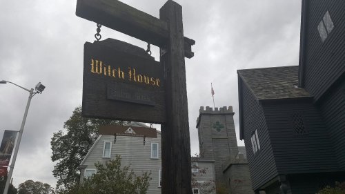 Sign for "Witch House" with a dark, cloudy sky and historic buildings in the background.