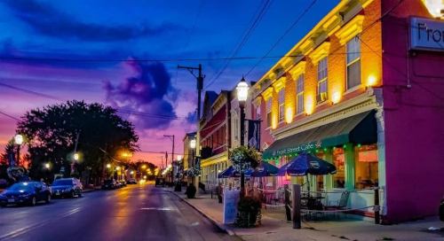 A vibrant sunset casts colorful hues over a charming street lined with cafes and shops, illuminated by warm lights.