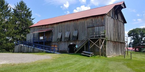 A large wooden barn with a red roof, surrounded by green grass and trees under a blue sky with clouds.