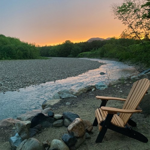 A serene riverside scene at sunset, featuring a wooden chair by a fire pit and a calm, winding river.