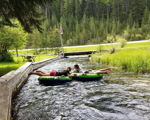 Two children float on colorful inner tubes in a serene river, surrounded by lush greenery and trees.