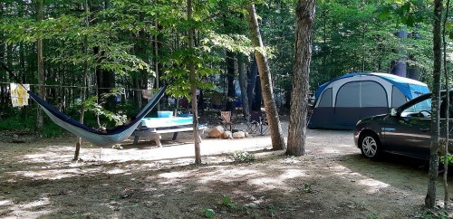 A campsite in a forest with a tent, hammock, picnic table, and a car parked nearby. Trees surround the area.