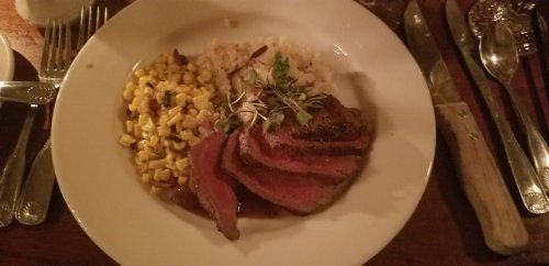 A plate of sliced steak with rice and corn, garnished with microgreens, set on a wooden table with utensils.