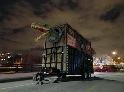 A large trailer with a dinosaur head protruding from the side, set against a night sky with clouds.