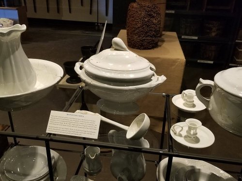 A display of white porcelain tableware, including a covered dish, teapot, and small cups, in a museum setting.