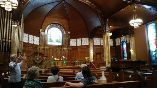 Interior of a church with wooden pews, stained glass windows, and a speaker gesturing towards the altar.