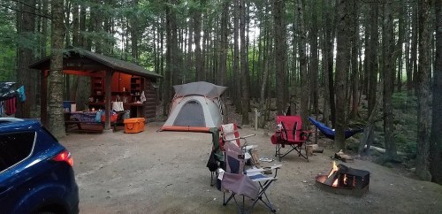 A campsite in a forest with a tent, chairs, a fire pit, and a picnic shelter surrounded by trees.