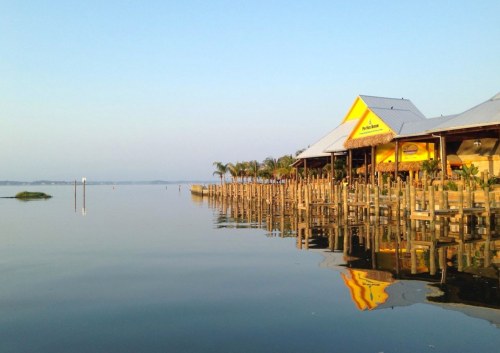 A calm waterfront scene with a yellow building reflecting on the water, surrounded by palm trees and wooden piers.