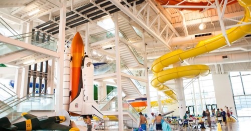 Indoor water park featuring a large rocket structure and colorful water slides, with people enjoying the space.