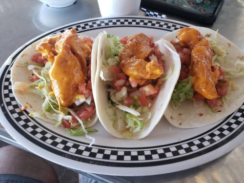 Three tacos filled with shrimp, lettuce, and tomatoes, drizzled with sauce, served on a checkered plate.