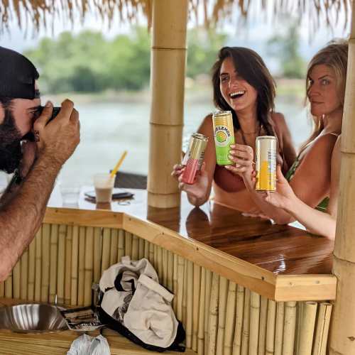 Three women hold colorful drinks while posing for a photo at a tiki bar, with a man taking their picture.
