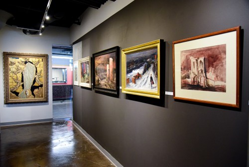 A gallery wall displaying various framed paintings in an art exhibition space.