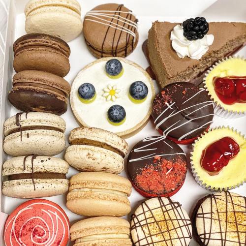 A variety of colorful macarons and desserts, including a chocolate cake slice and fruit-topped treats, arranged in a box.