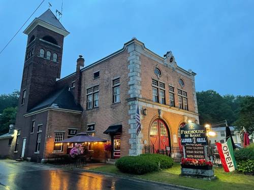 Historic firehouse building at dusk, featuring a restaurant sign and outdoor seating under a cloudy sky.