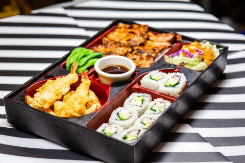 A bento box featuring shrimp tempura, grilled chicken, sushi rolls, salad, and dipping sauce on a striped table.