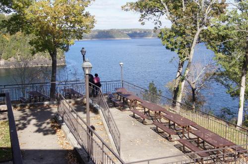 A serene lakeside view with picnic tables and two people sitting by the water, surrounded by trees and autumn foliage.