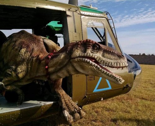 A dinosaur figure emerges from a military helicopter, set against a clear blue sky and grassy field.