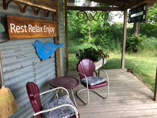 A cozy porch with two chairs, a small table, and a sign that says "Rest Relax Enjoy," surrounded by greenery.