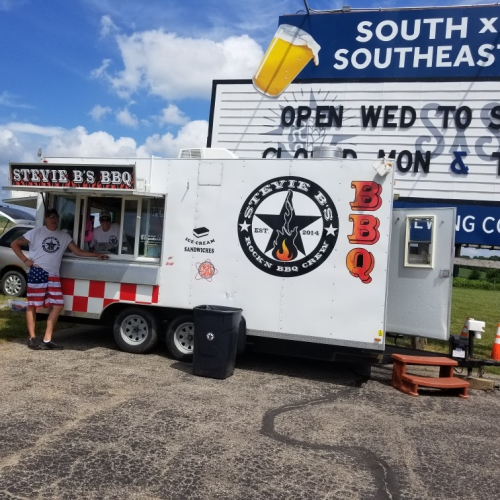 A food truck named "Stevie B's BBQ" with a menu sign, parked in a sunny outdoor area. People are ordering food.