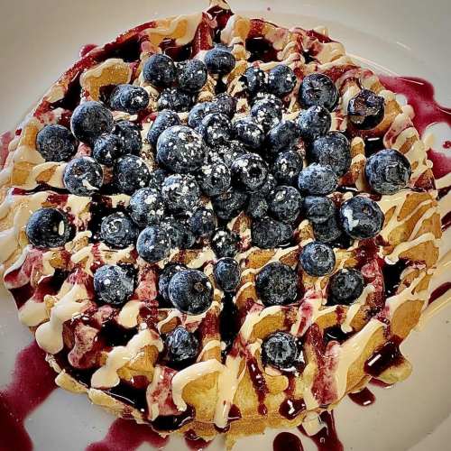 A delicious waffle topped with fresh blueberries, drizzled with berry sauce and cream, sprinkled with powdered sugar.