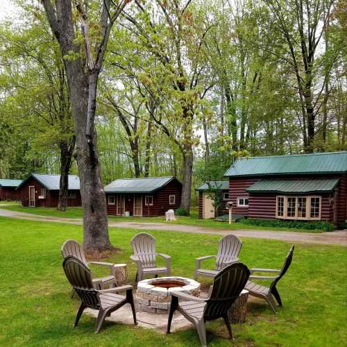 A cozy outdoor scene with a fire pit surrounded by chairs, nestled among trees and rustic cabins.