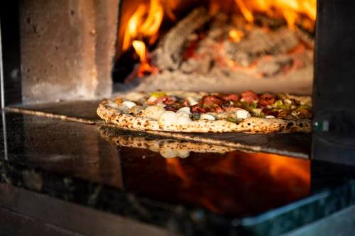 A freshly baked pizza with toppings, placed in a wood-fired oven with flames in the background.