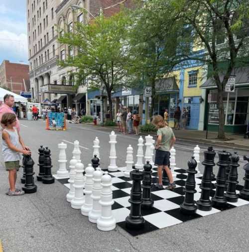 Two children play on a large outdoor chessboard with oversized black and white pieces in a street setting.