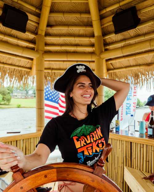 A smiling woman in a pirate hat stands at the helm of a boat, surrounded by tropical decor and flags.