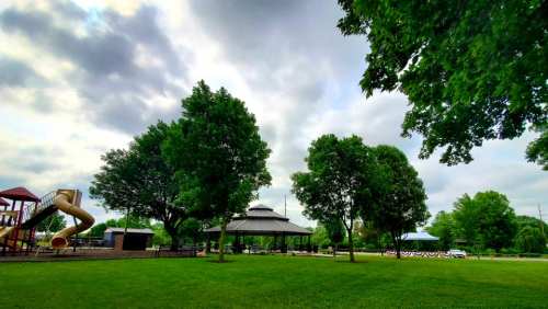 A park scene featuring a playground slide, a gazebo, and lush green trees under a cloudy sky.
