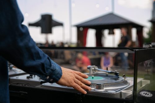 A DJ's hand adjusts a vinyl record on a turntable at an outdoor event, with people in the background enjoying the music.