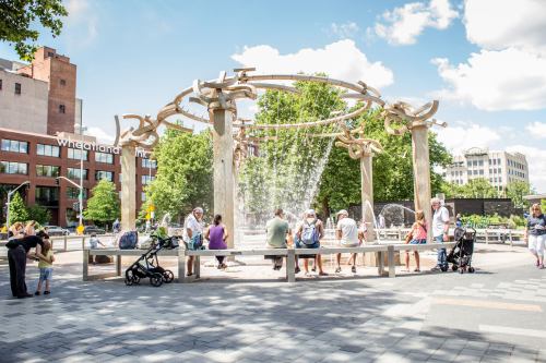 A lively park scene with a fountain, people gathered around, and greenery under a bright blue sky.