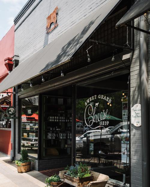 Exterior of Sweet Grass Cheese Shop, featuring a storefront with large windows and potted plants, on a sunny day.