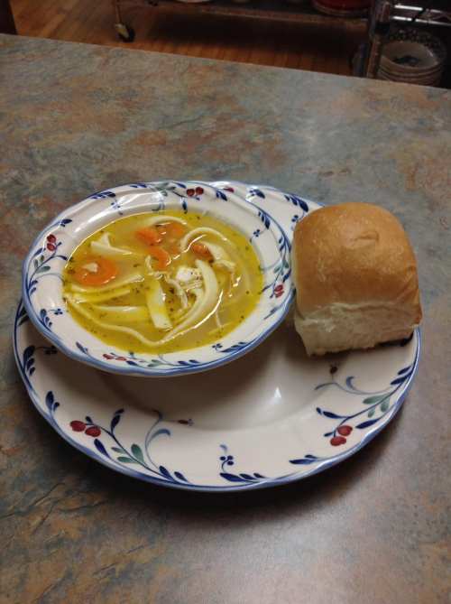 A bowl of chicken noodle soup with carrots and a soft dinner roll on a decorative plate.