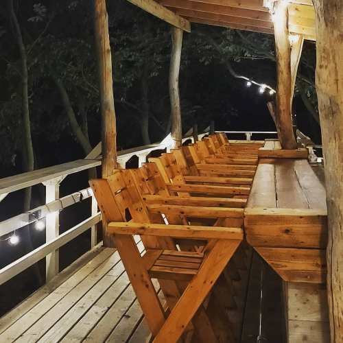 A wooden deck at night with a long table and several chairs, illuminated by soft lights and surrounded by trees.