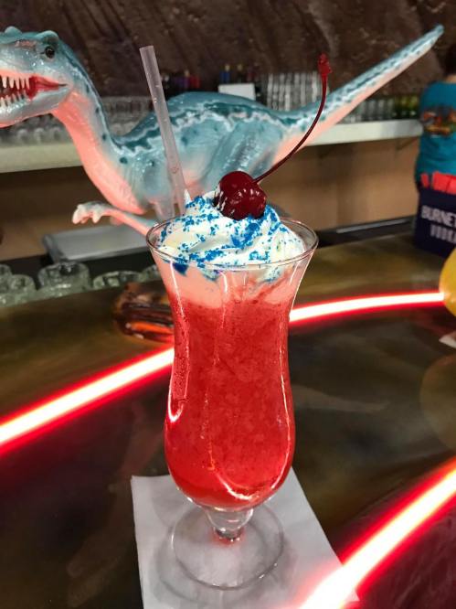 A vibrant red cocktail topped with whipped cream, blue sprinkles, and a cherry, served in a tall glass at a bar.